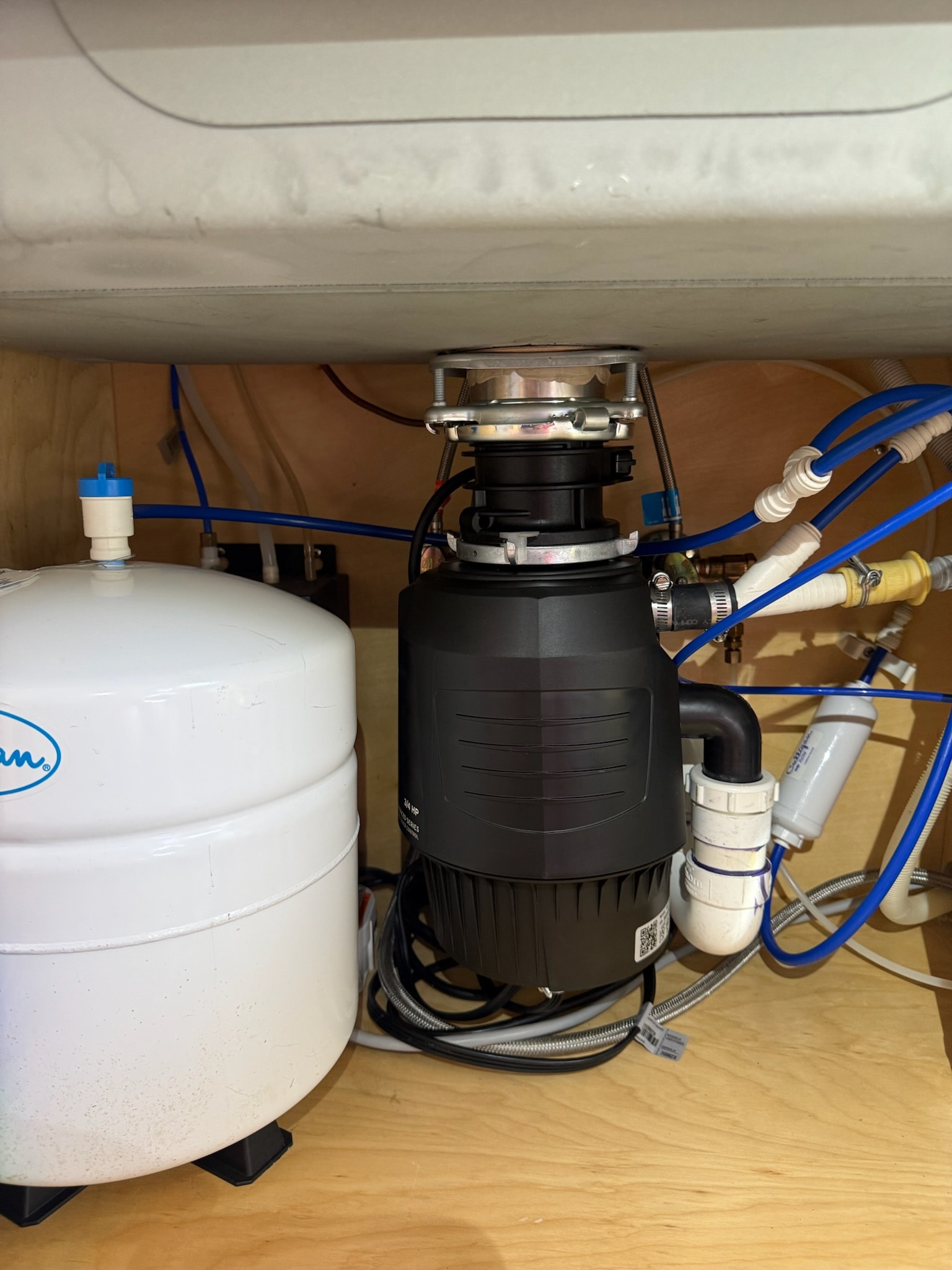 A 3/4 horsepower garbage disposal installed under a kitchen sink, beside a drinking-water tank, with the trap and supply lines connected