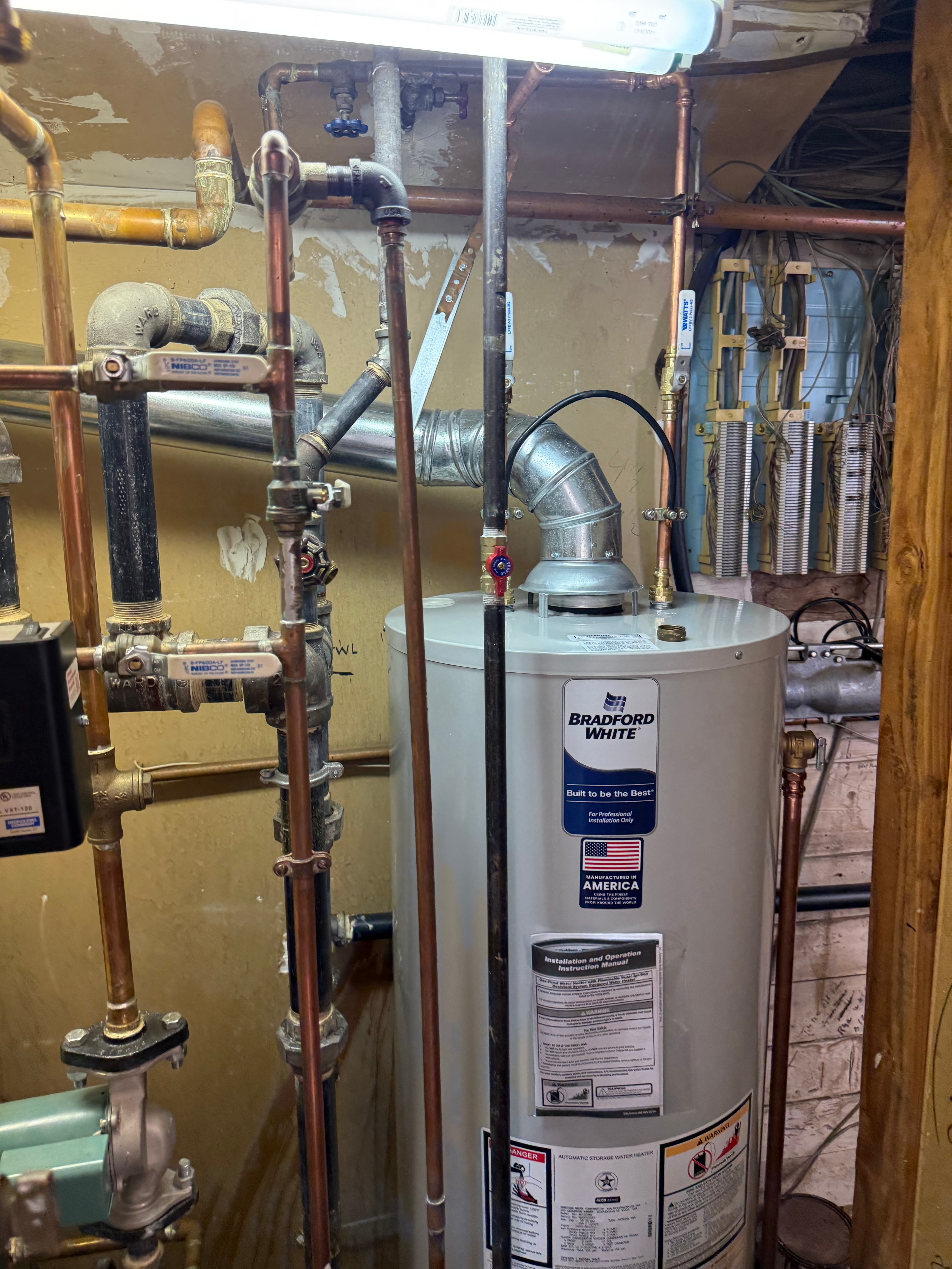 A Bradford White gas water heater newly fitted beside existing boiler piping in a basement